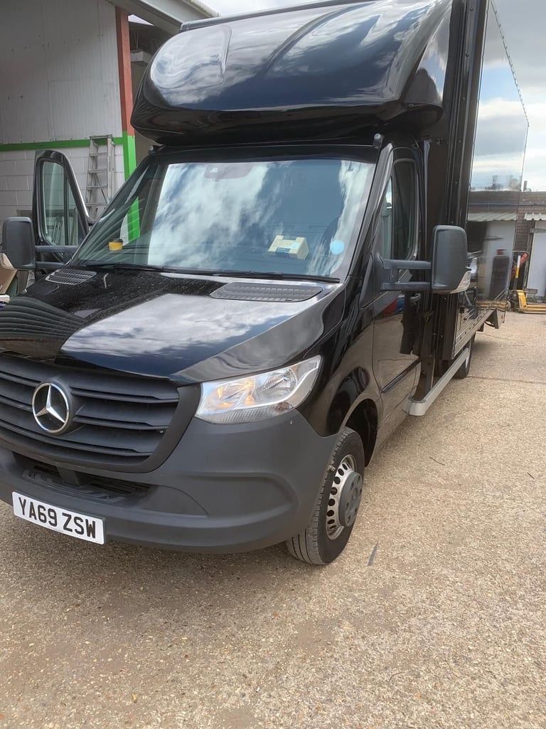 Black Mercedes-Benz campervan with open roof hatch and windscreen cover parked on asphalt lot
