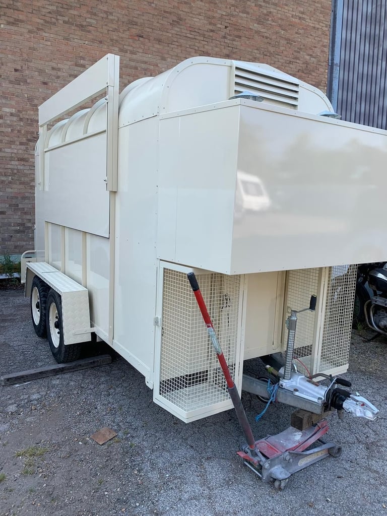 White two-horse trailer with refrigeration unit parked against brick building, ramp deployed