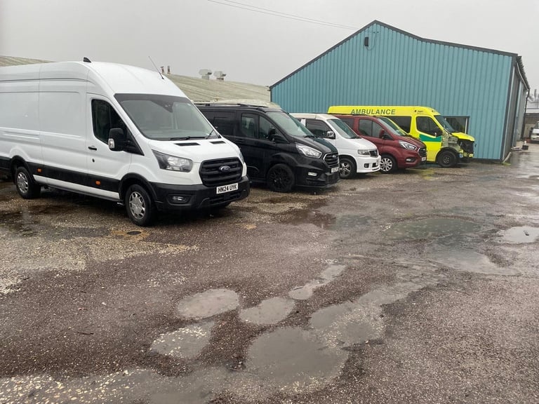 Row of parked vehicles including white van, black car, and yellow emergency vehicle in front of turquoise industrial building